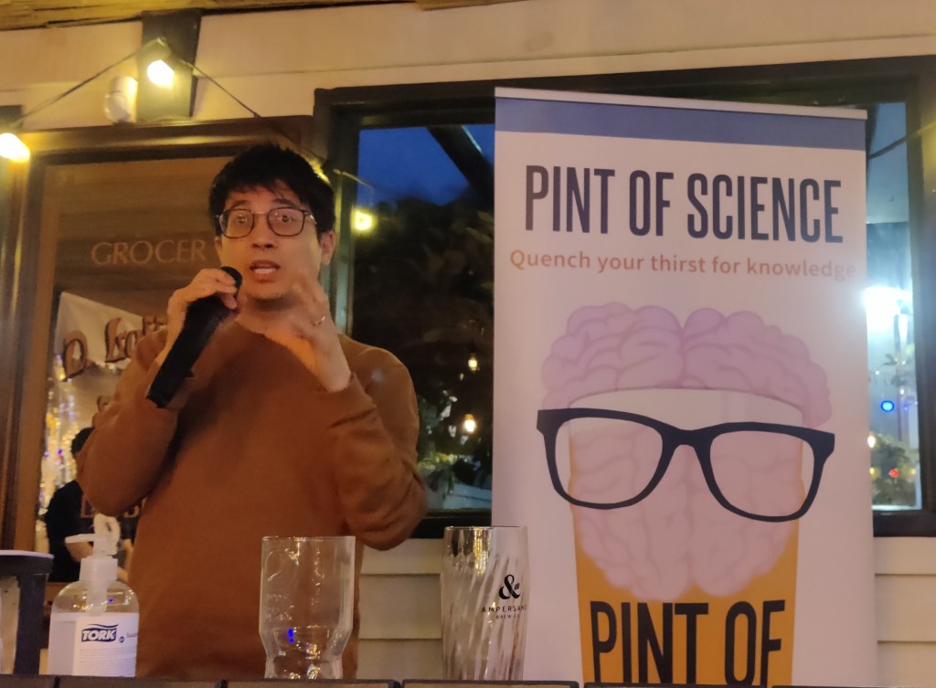 Dark-haired man with glasses and a brown jumper speaking into a microphone, standing next to a roller banner with the Pint of Science festival writing and logo of a brain wearing glasses on top of a glass of yellow liquid.