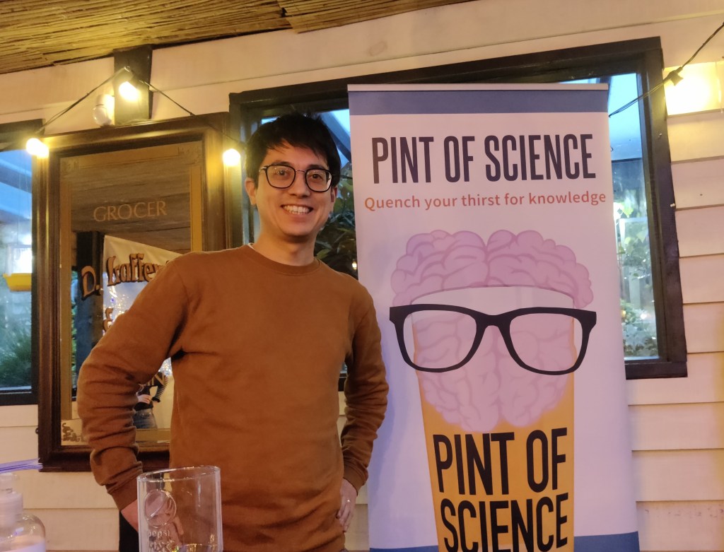 Dark-haired man with glasses and a brown jumper standing smiling next to a roller banner with the Pint of Science festival writing and logo of a brain wearing glasses on top of a glass of yellow liquid.